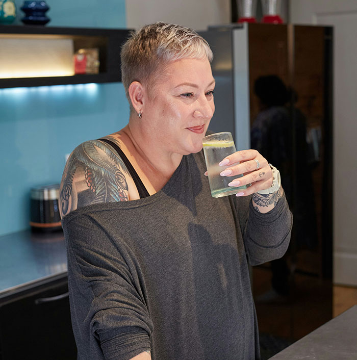 Person with a short hairstyle and tattoos enjoying a drink in a kitchen, possibly sharing dark secrets.