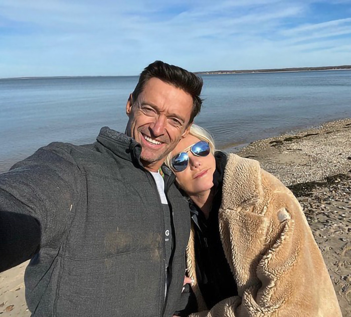 Deborra-Lee Furness and Hugh Jackman smiling on a beach, wrapped in a cozy coat.