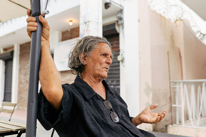 Man sitting on an outdoor porch, engaged in conversation, wearing a black shirt with glasses hanging from it.