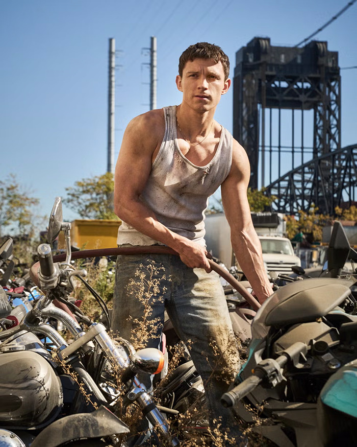 Tom Holland in a white tank top surrounded by motorcycles, urban backdrop, embodying a rugged style.