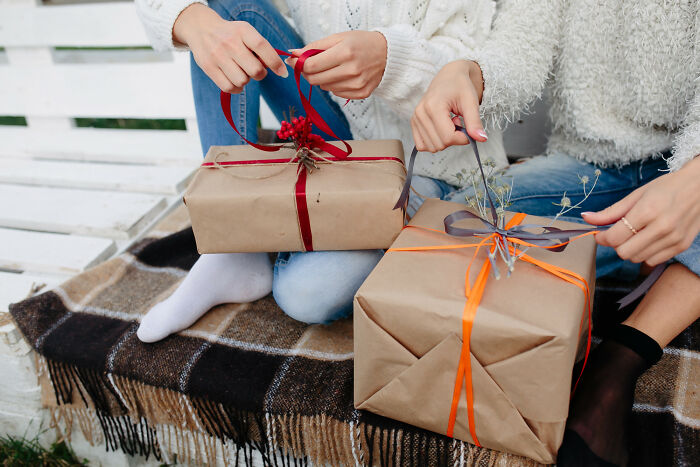 Two people sitting on a bench, unwrapping Christmas gifts wrapped in brown paper with red and orange ribbons.