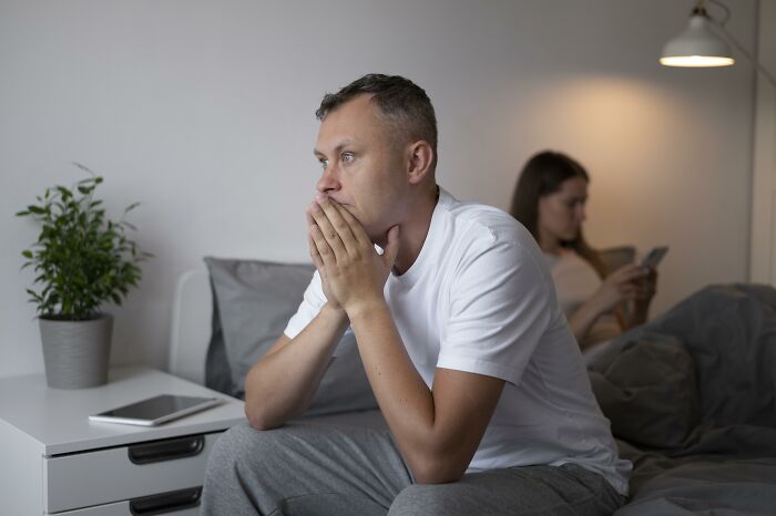 Man in deep thought sitting on bed, woman using phone in the background, reflecting an unreasonable friend relationship. Man in deep thought sitting on bed, woman using phone in the background, reflecting an unreasonable friend relationship.