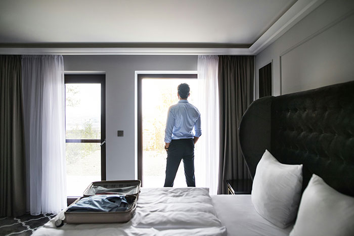Man in hotel room looking out the window with open suitcase on bed, following hotel rules.