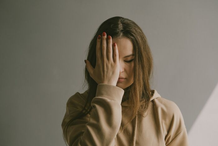 Teen girl frustrated with parents, hand on face, wearing a beige hoodie against a neutral background.