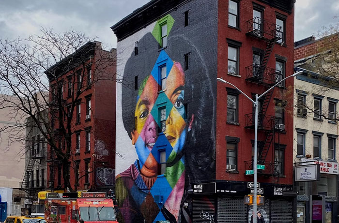 Colorful mural on a building facade in city, featuring faces merged in geometric patterns, surrounded by urban street scene.