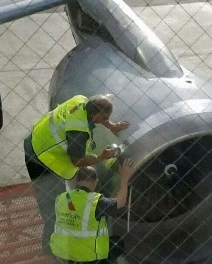 Workers in yellow vests inspecting a jet engine through a fence, capturing a bizarre and funny moment.