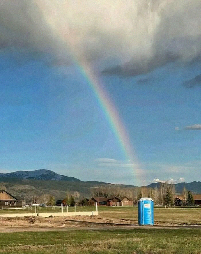 A bizarre and funny scene of a rainbow ending at a portable toilet in a field.