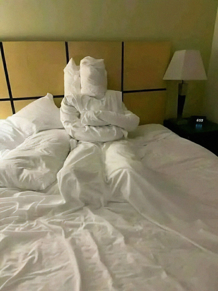 Pillows arranged humorously on a bed, resembling a sitting figure, creating a funny scene in a room.