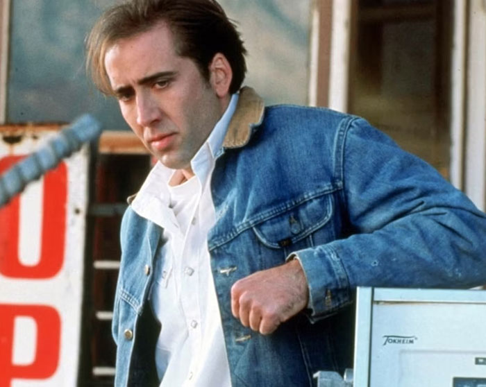 Man in denim jacket leaning on a railing, outdoor setting, rich people rumors hearable in taxi rides.