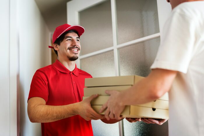 Delivery person in a red uniform smiling while handing over pizza boxes at a door.
