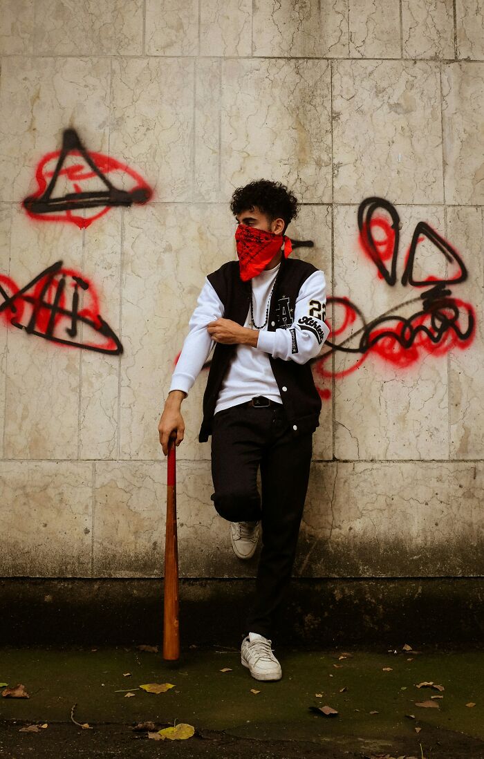 Person in a red bandana and varsity jacket leaning against graffiti-covered wall, holding a bat. Awkward delivery scenario.
