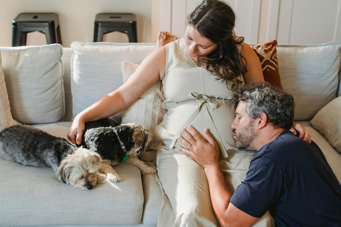 Pregnant woman in white dress sits on sofa with partner, embracing her belly, surrounded by two small dogs.