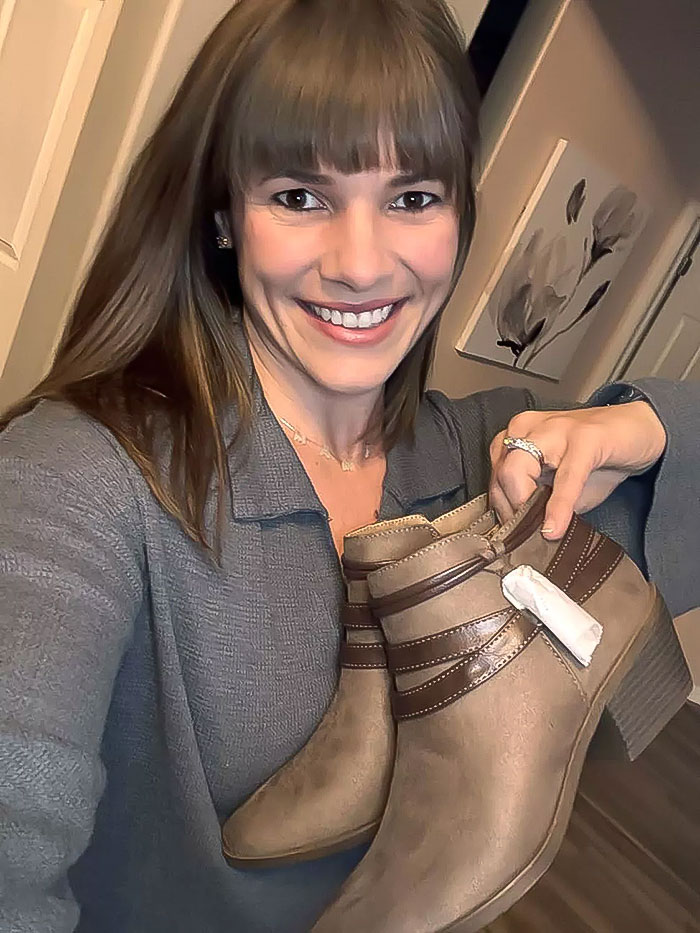 Woman smiling and holding thrifted boots with tags, representing a gift for her MIL. Woman smiling and holding thrifted boots with tags, representing a gift for her MIL.