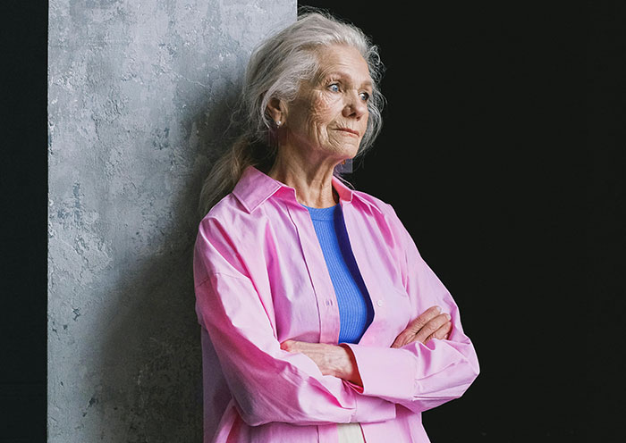 Elderly woman in a pink shirt looks thoughtful, arms crossed, against a grey wall. Elderly woman in a pink shirt looks thoughtful, arms crossed, against a grey wall.