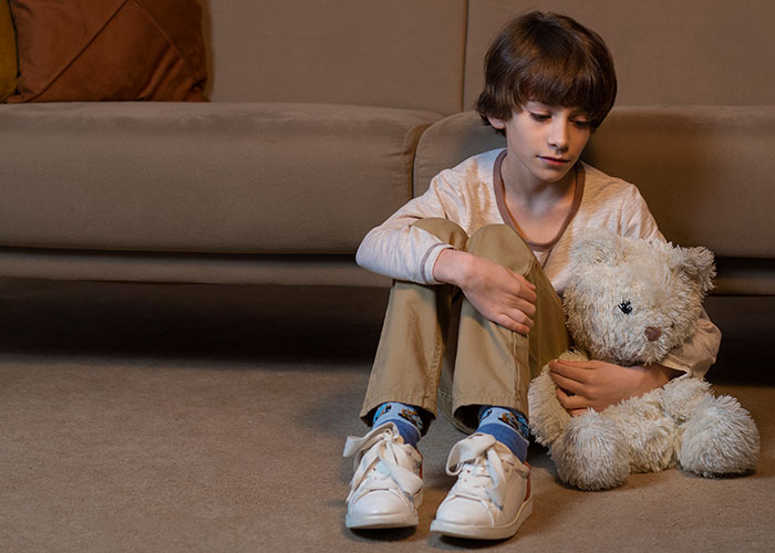 A child sitting on the floor hugging a teddy bear, looking contemplative, next to a sofa.