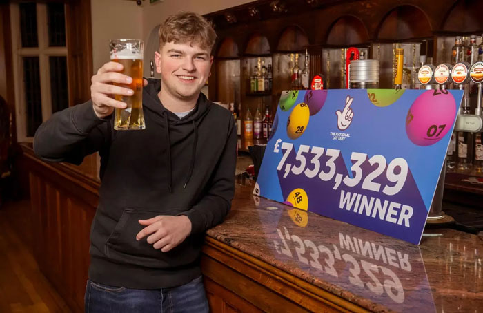 Man in a pub celebrating a &pound;7.5M lottery win with a pint, standing next to a winning sign.