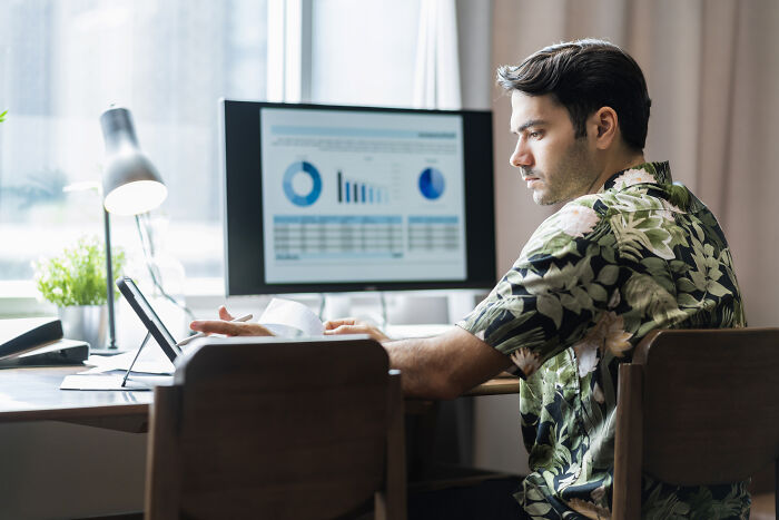 Man in floral shirt working remotely at a desk, analyzing charts on a computer screen.