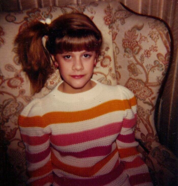 Young girl in an 80s fashion striped sweater, sitting on a patterned chair.