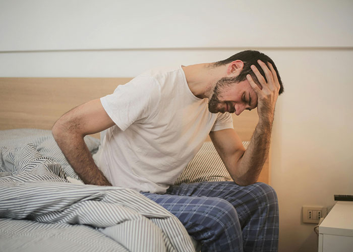 Man in pajamas sitting on bed holding head, illustrating a stressful American healthcare experience.