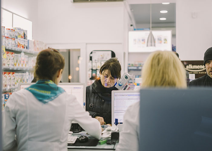 Pharmacy counter scene with staff assisting customers, reflecting challenges in American healthcare.