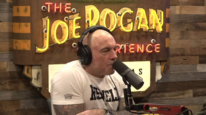 A comedian speaking into a microphone on a podcast set, wearing headphones and a t-shirt.