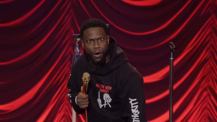 A comedian on stage with a surprised expression, wearing a black hoodie and holding a golden microphone.