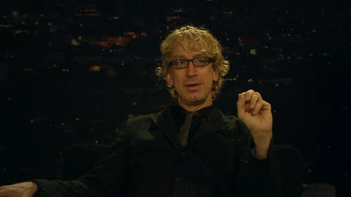A comedian in a black suit and glasses, sitting against a dark background, gesturing with one hand.