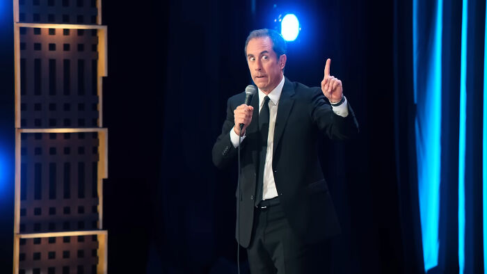 Comedian performing on stage with a microphone, wearing a suit, under a spotlight.