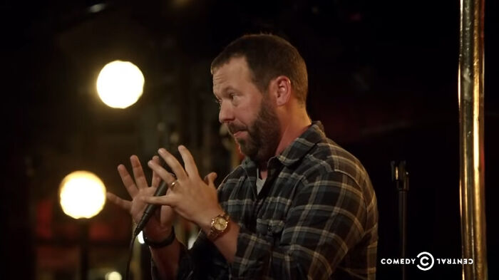 Comedian on stage with microphone in hand, wearing a plaid shirt, performing in a dimly lit comedy club setting.
