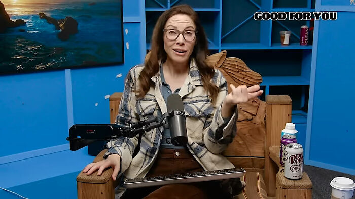 Comedian in a checkered jacket during a podcast session, surrounded by a microphone and beverages.