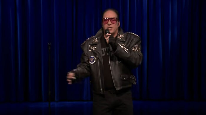 Comedian in a studded leather jacket and red glasses performing on stage, blue curtain backdrop.