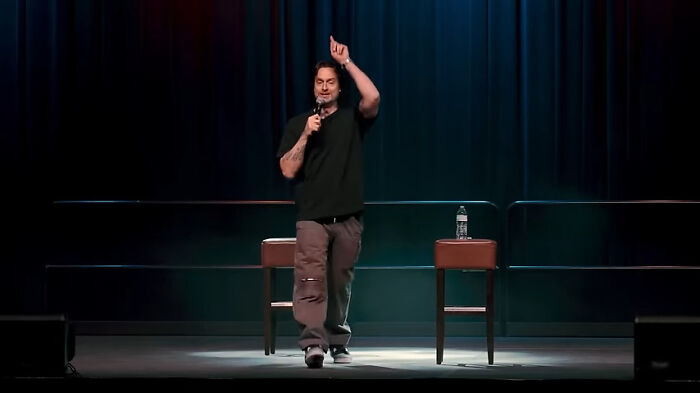 Comedian performing on stage with microphone, a chair, and water bottle.