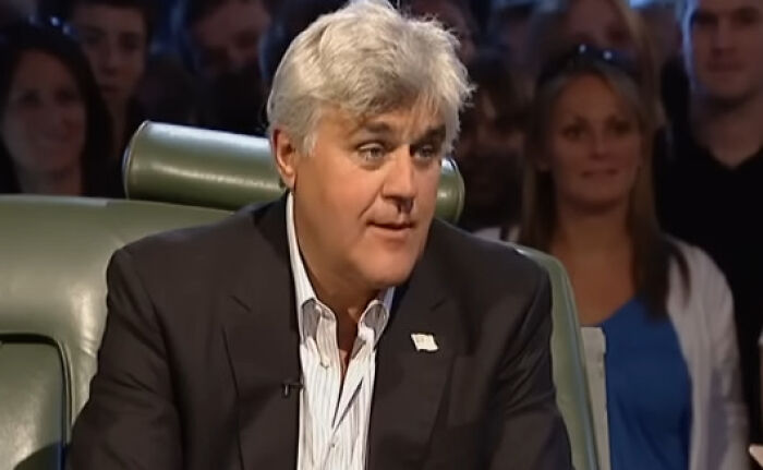 A comedian on a talk show, wearing a suit, discussing mainstream success with an attentive audience in the background.