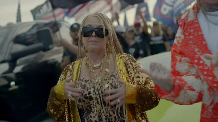 Person wearing sunglasses and a gold sequin jacket at an outdoor event, surrounded by people and flags.