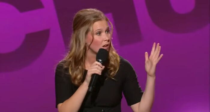 Female comedian performing on stage, holding a microphone, with a purple background.