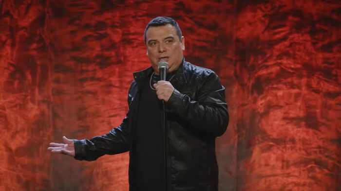 Comedian performing on stage with a red background, holding a microphone, and gesturing with one hand.