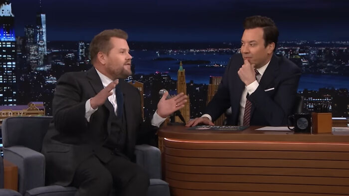 Two comedians in suits talking during a late-night TV show, featuring city skyline in the background.