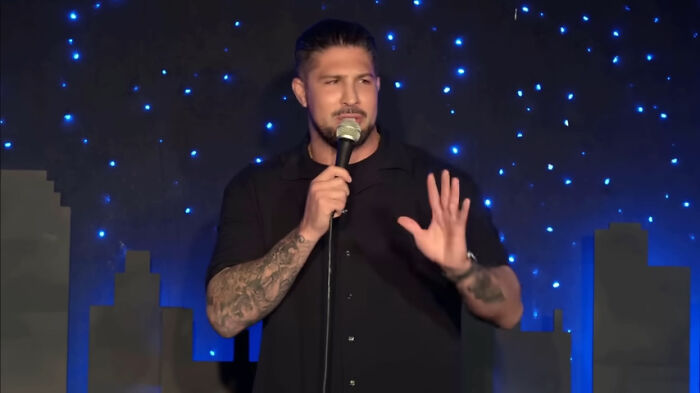 Comedian performing on stage, holding a microphone, with a cityscape backdrop and blue sparkly lights.