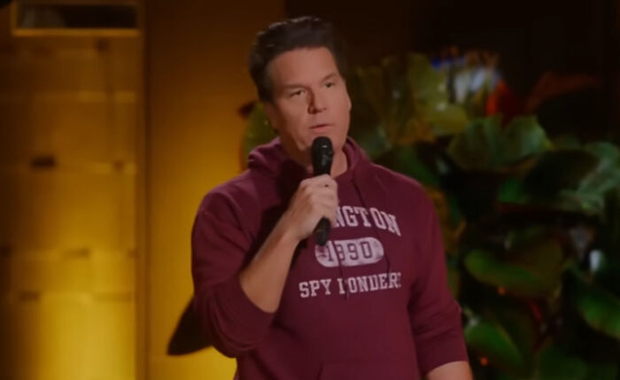 A comedian in a maroon hoodie performs on stage, holding a microphone, with plants in the background.
