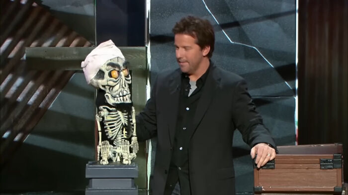 Comedian with a skeleton puppet on stage, performing mainstream comedy act.