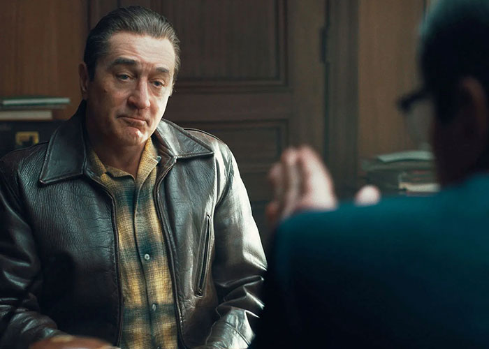 Man in a leather jacket sitting at a table in a film scene, possibly discussing casting choices in Hollywood.