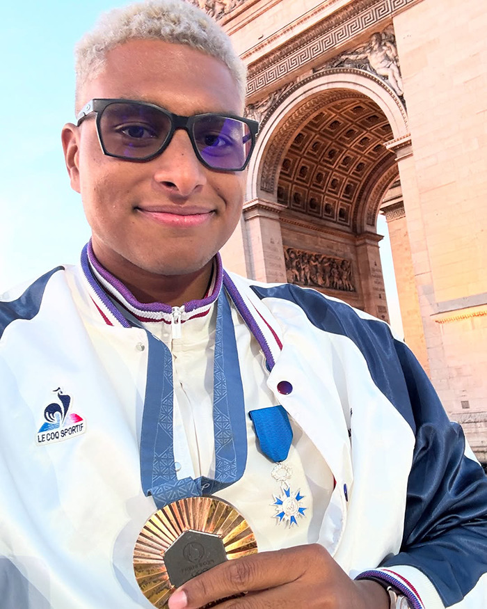 Athlete in Paris holding a medal, standing near a historic arch, wearing dark glasses and a sports jacket.