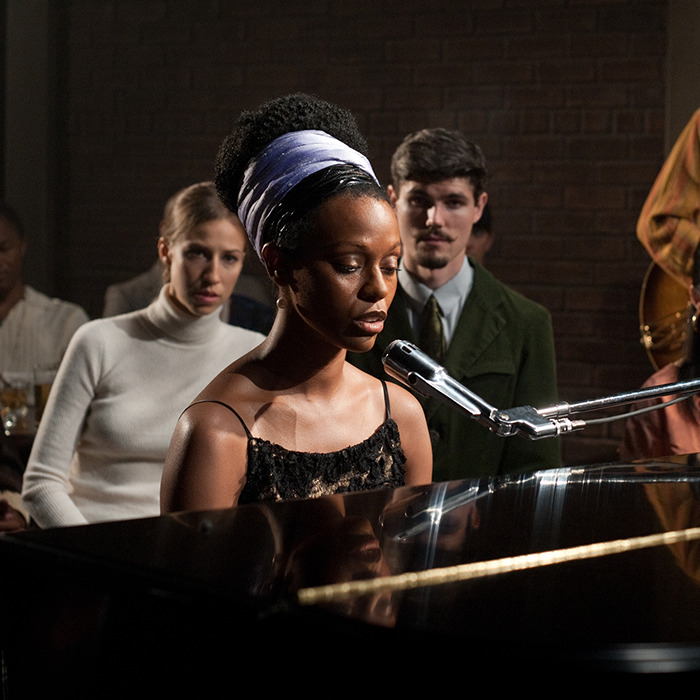A woman with a headband sings at a piano, surrounded by an attentive audience. A woman with a headband sings at a piano, surrounded by an attentive audience.