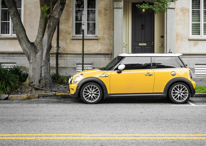 A yellow Mini Cooper parked on a street next to a tree and building.