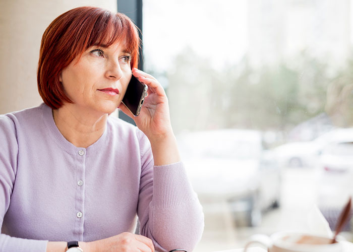 Woman with red hair in a lavender sweater, talking on the phone, sitting by a window, possibly contemplating an influencer request.