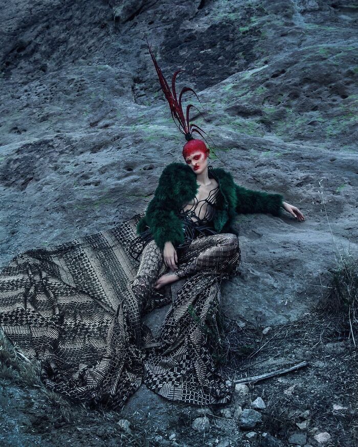 Unique model in elaborate fashion with a red headpiece and green fur coat, posed against a rocky background.