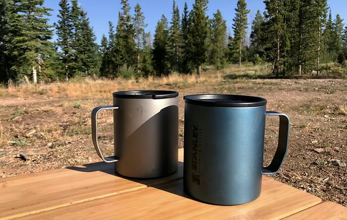 Two insulated mugs on a picnic table in a forest, ideal gift ideas for someone who has everything.