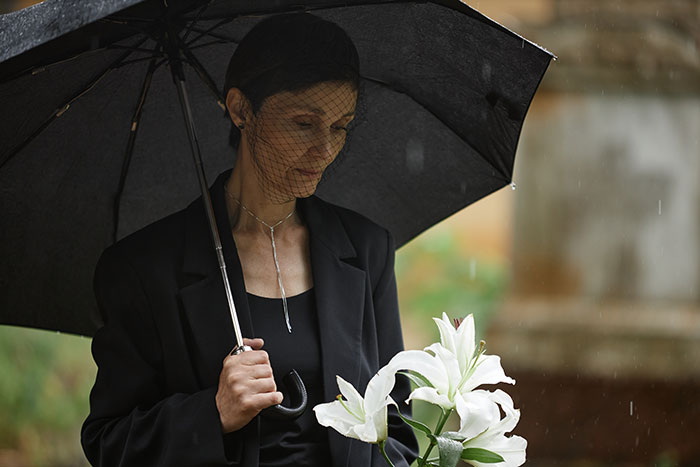 A woman in mourning attire holds lilies and an umbrella at a funeral, related to inheritance issue.