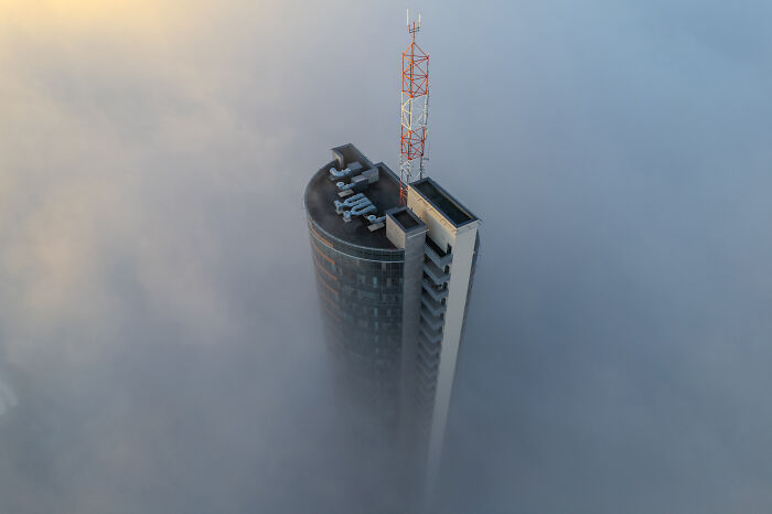 Lithuania bird's-eye view shows a tall building emerging through mist and clouds at dawn.