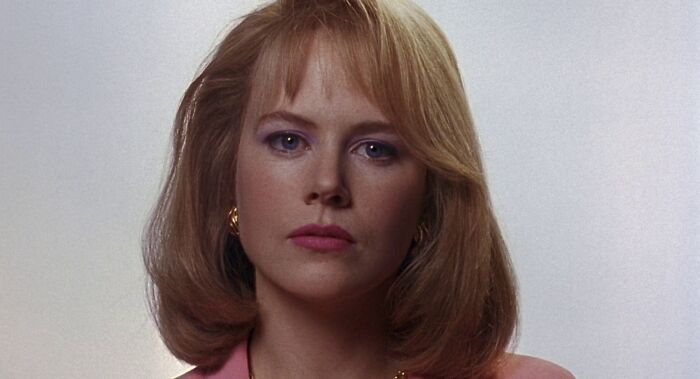 Blonde woman in a pink jacket, reminiscent of Celebrities-In-The-90s, with a serious expression against a plain background.